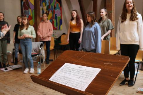 eine Gruppe Sängerinnen der Young Voices Brandenburg ist bei der Probe zu sehen, im Vordergrund sieht man die Noten des Songs "What Would You Do?"